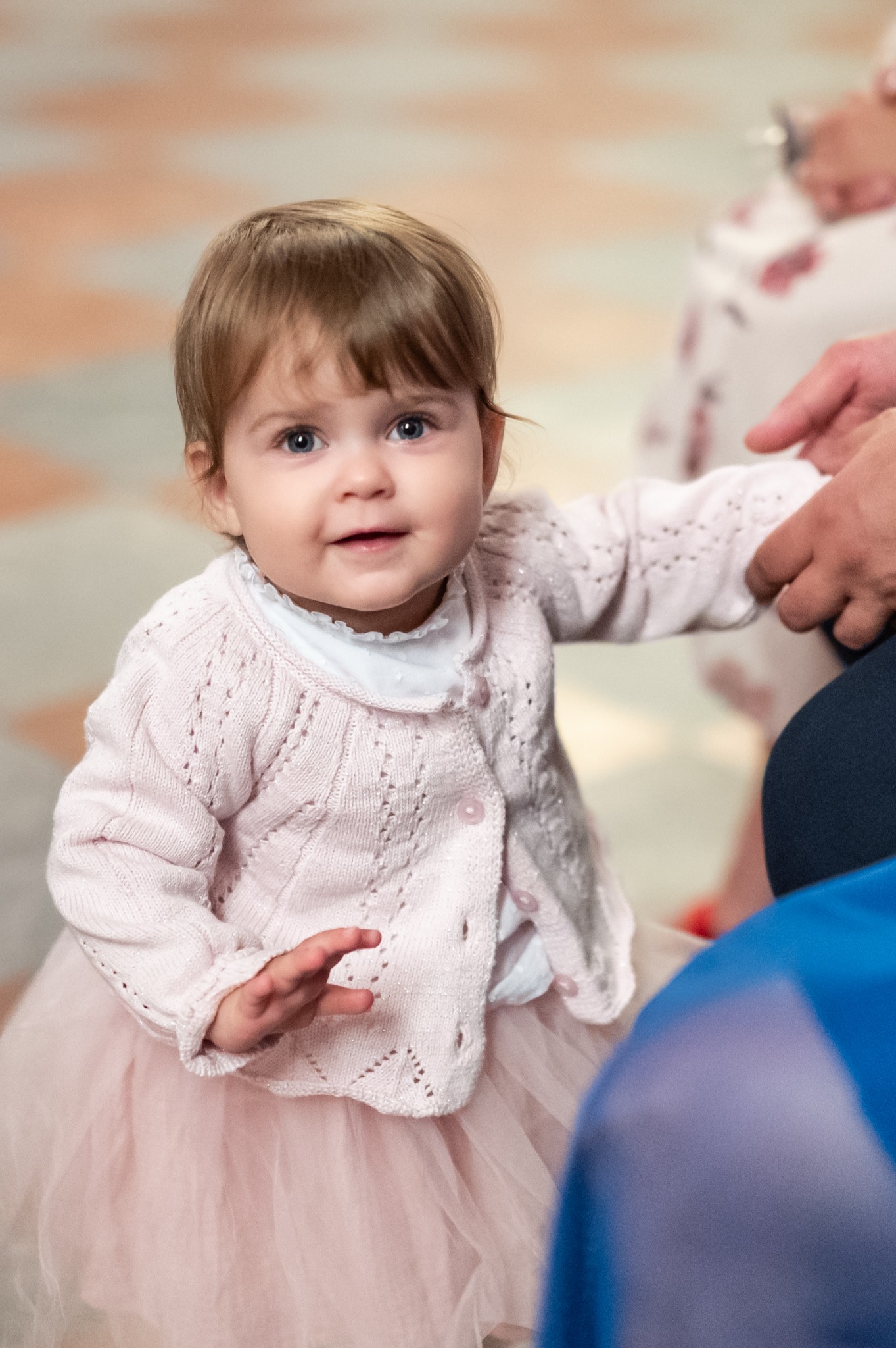 Urocza dziewczynka w różowej sukience i sweterku, trzymana za rękę przez dorosłego. Jasne, naturalne światło, miękkie tło. Ujęcie z dołu, podkreślające dziecięcą niewinność.