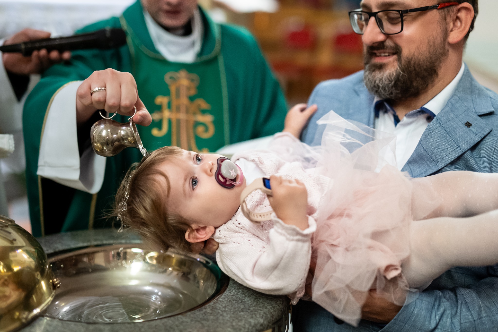 Chrzest dziewczynki w różowej sukience, trzymanej przez ojca w niebieskiej marynarce. Ksiądz polewa wodą z naczynia. Ujęcie z bliska, skupione na dziecku i ceremonii.