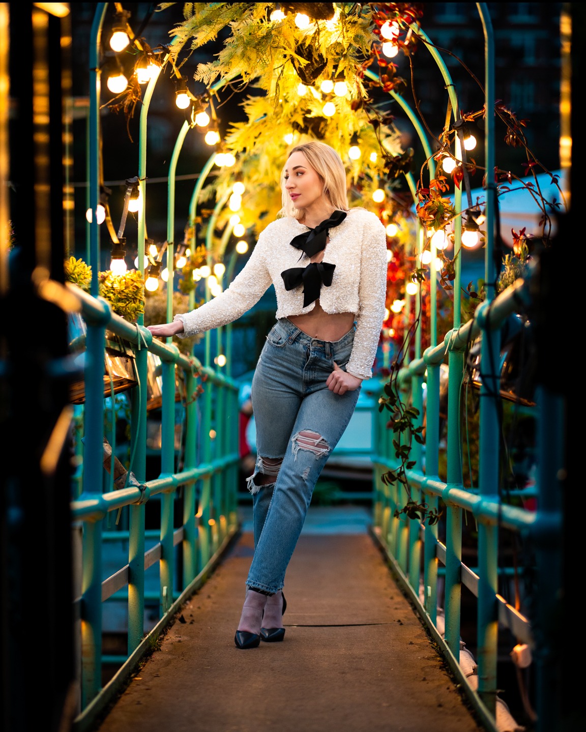 Blond modelka w dżinsach i białym swetrze z kokardami pozuje na moście z lampkami, odwrócona w lewo, z nogą wysuniętą do przodu. Ujęcie w ciepłych barwach.