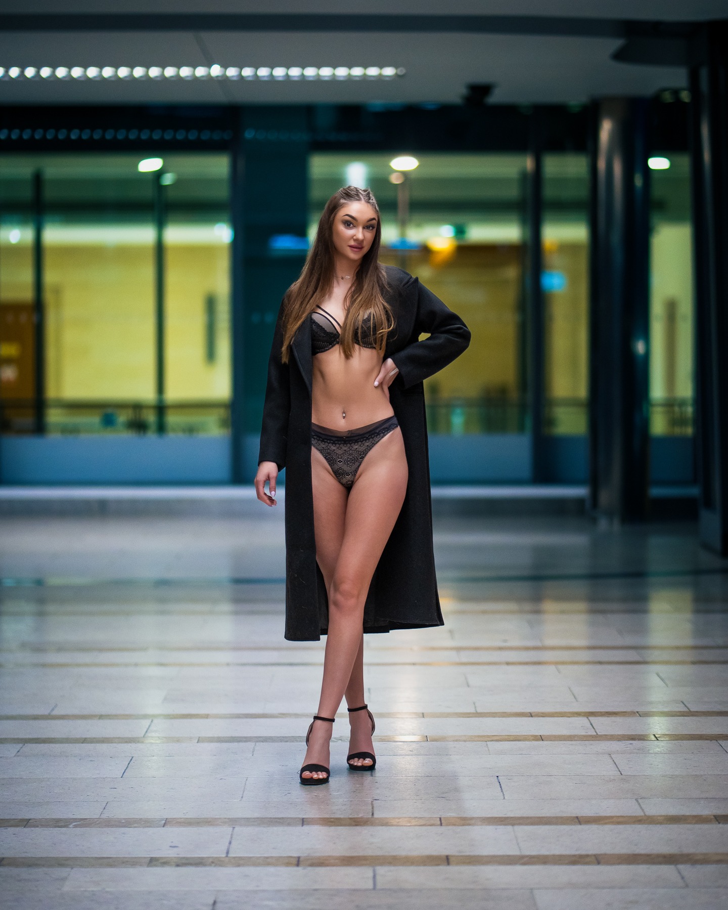 Kobieta w bieliźnie i płaszczu pozuje na tle przeszklonej ściany. Ujęcie centralne, pełna sylwetka, wysokie obcasy, subtelny makijaż, długie włosy.