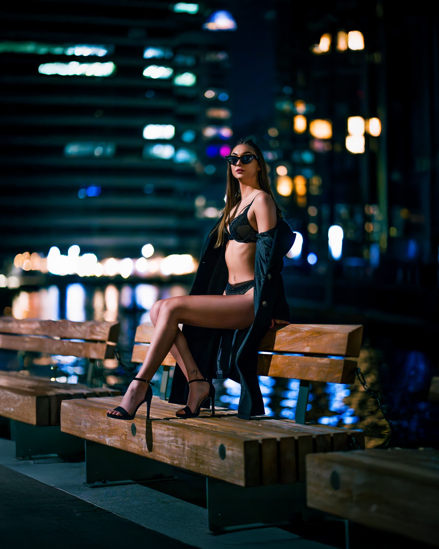 Kobieta w bieliźnie i płaszczu siedzi na ławce na tle nocnego miasta z rozmytymi światłami. Uwagę przykuwają wysokie obcasy i okulary przeciwsłoneczne.