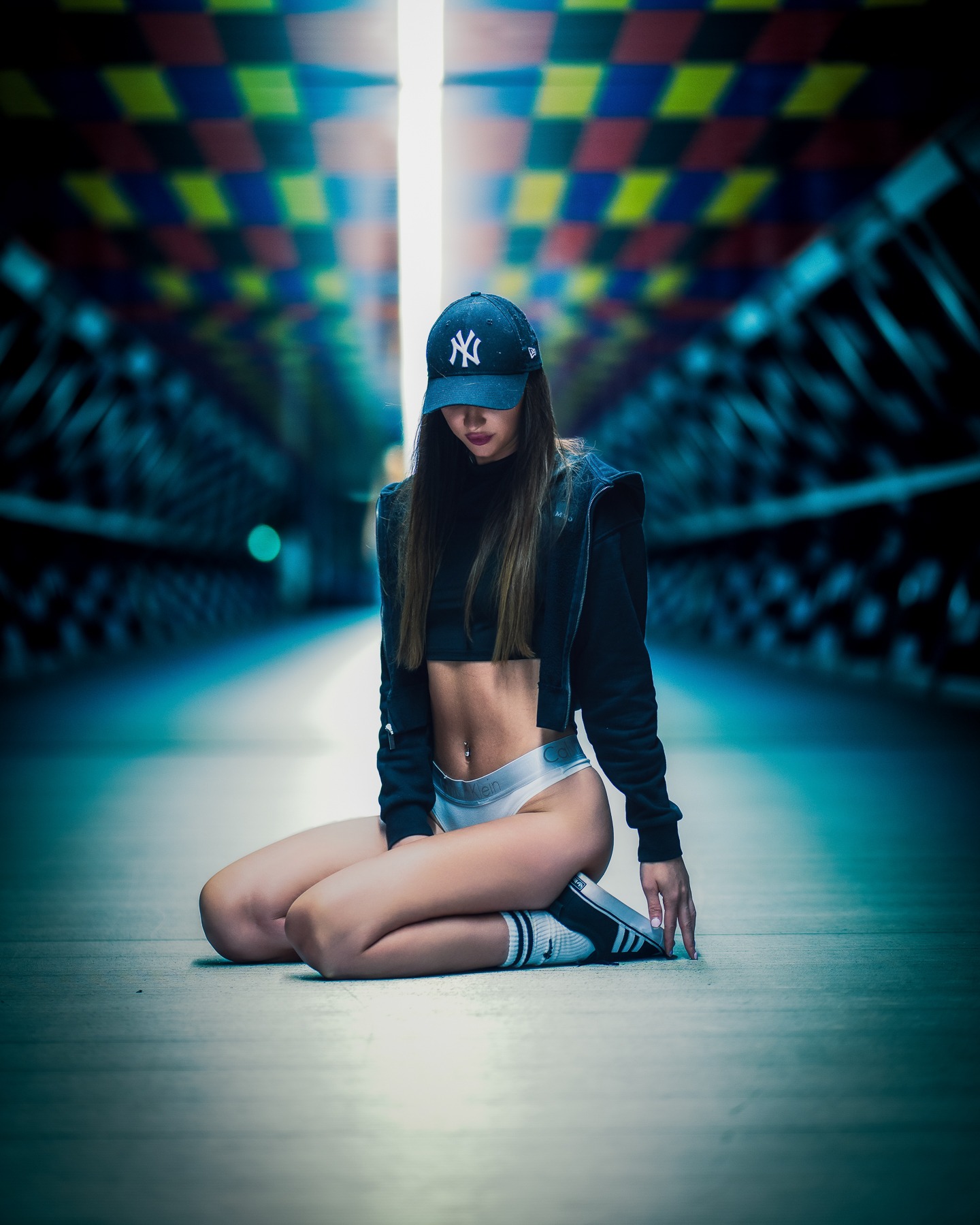 Kobieta w czapce z daszkiem klęczy w tunelu z kolorowym sufitem. Ubrana w sportowy top i bieliznę, pozuje w dynamicznym ujęciu z perspektywą.