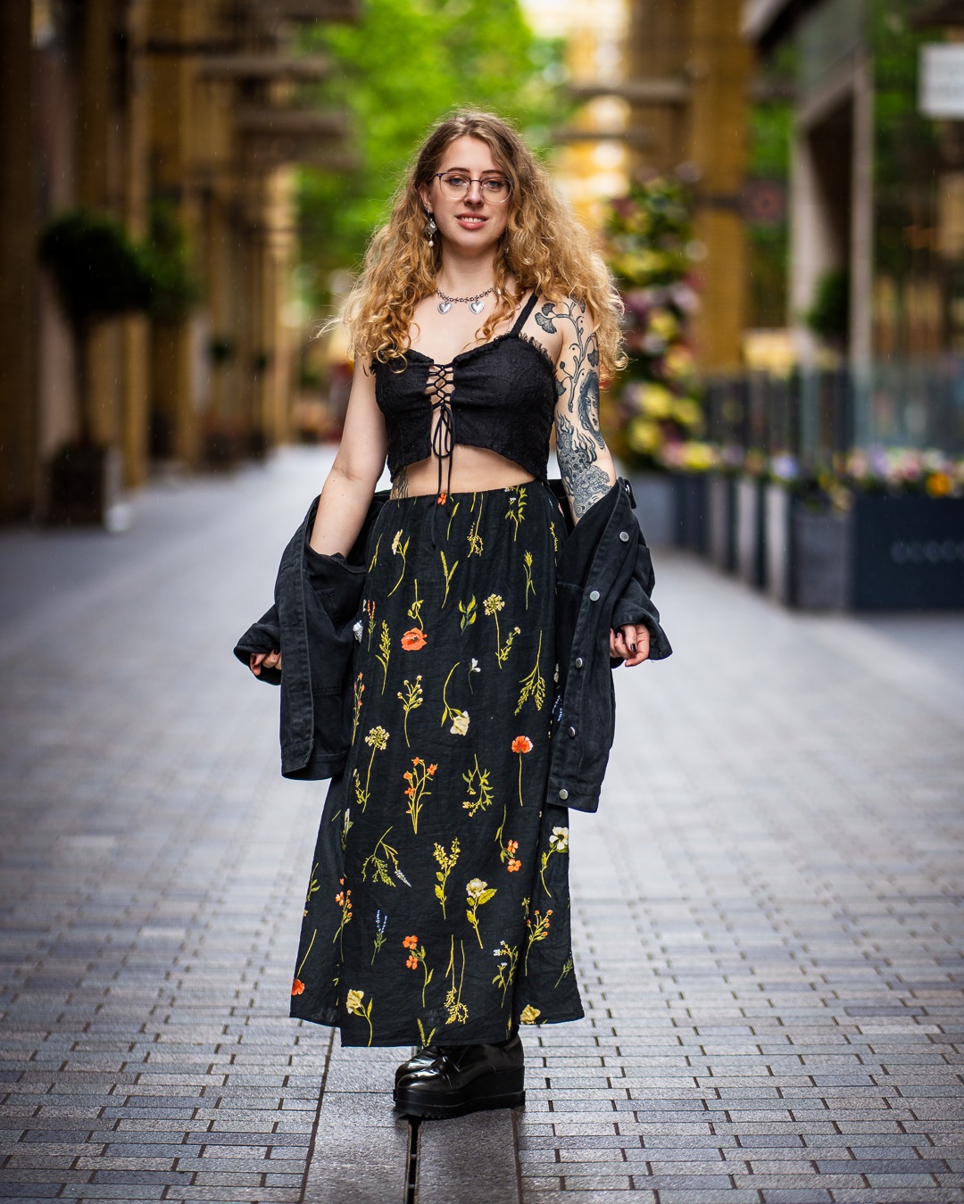 Portret kobiety w kwiecistej sukience i czarnym topie, stojącej na brukowanej ulicy z rozmytym tłem miejskim. Widoczne tatuaże na ramieniu i delikatna biżuteria.