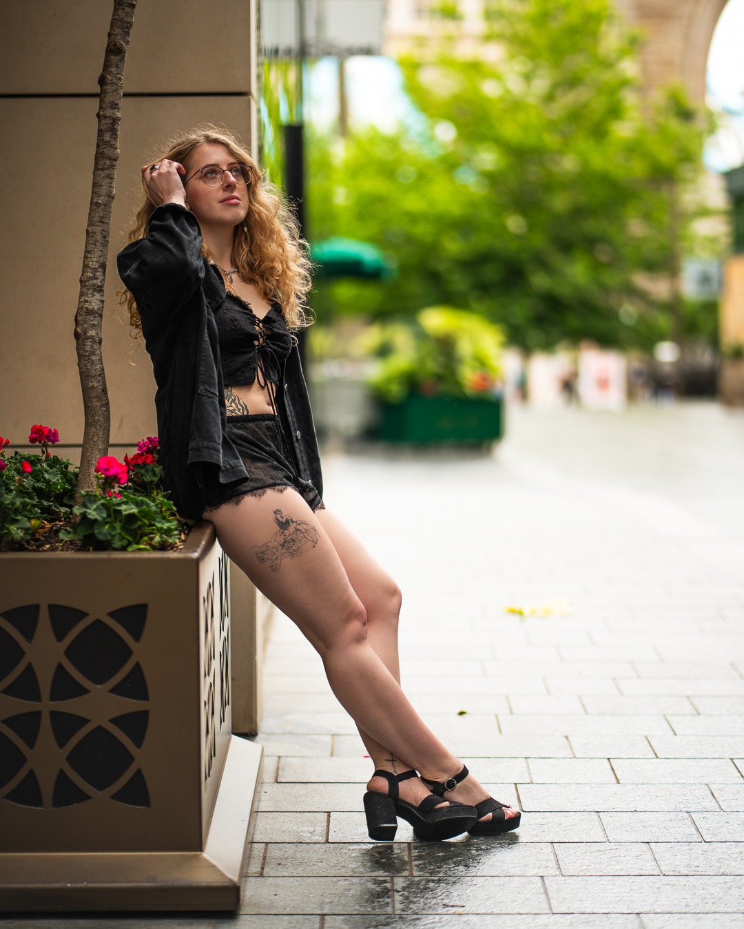 Portret kobiety w czarnym stroju, opierającej się o donicę z kwiatami na tle miejskiej uliczki. Uwagę przykuwają kręcone włosy i tatuaż na udzie.