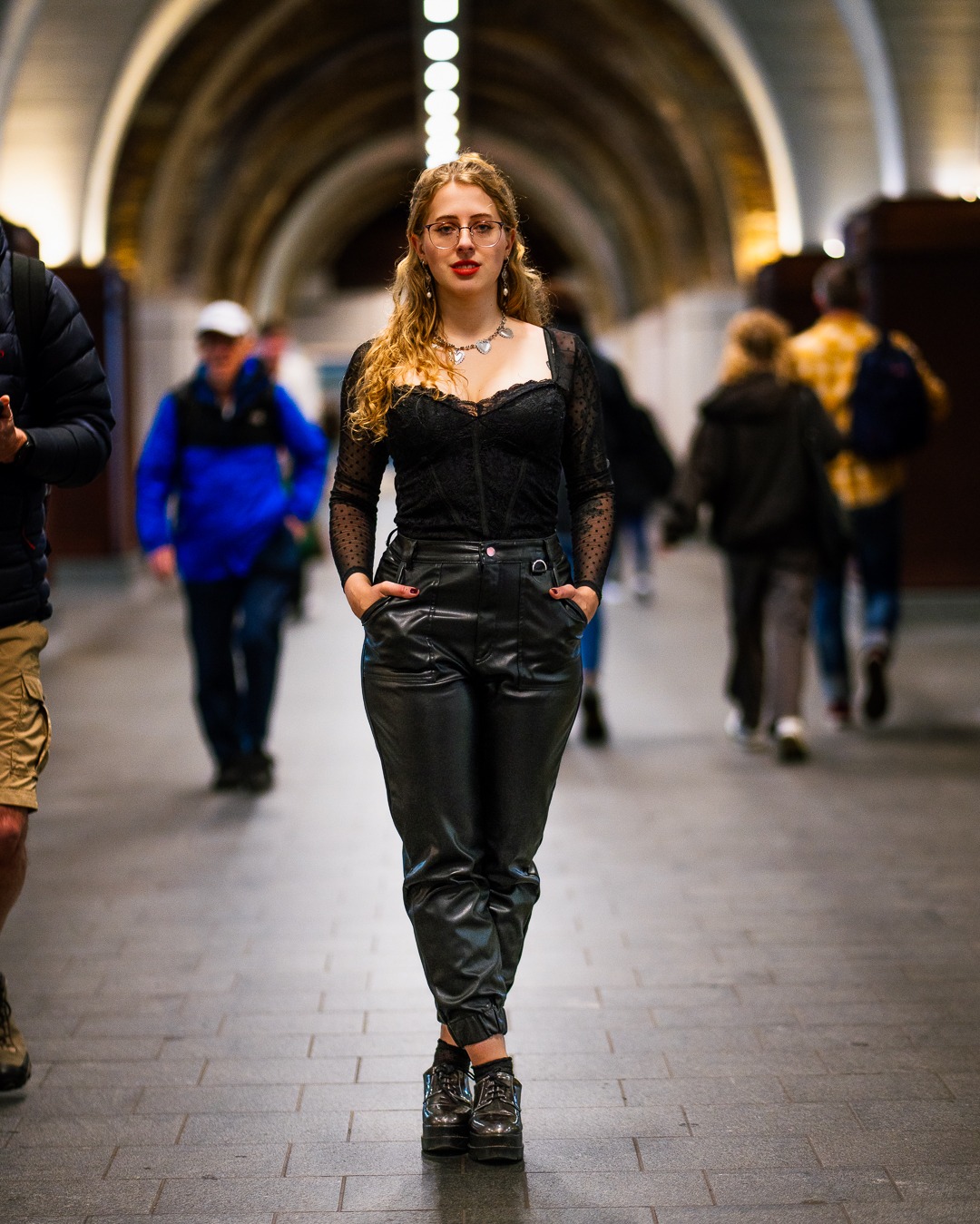 Kobieta w czarnym stroju pozuje w przejściu z łukowatym sufitem. Uwagę przyciągają okulary w czerwonych oprawkach i srebrny naszyjnik. Tło stanowią rozmyte sylwetki przechodniów.