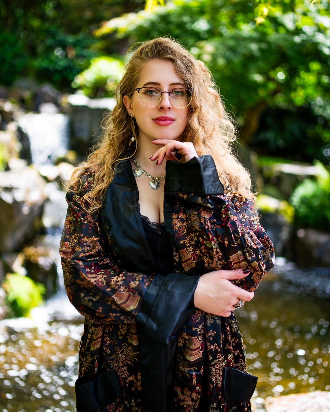 Portret kobiety w kimonie na tle wodospadu w japońskim ogrodzie. Modelka w okularach, z kręconymi włosami i biżuterią pozuje, opierając brodę na dłoni.