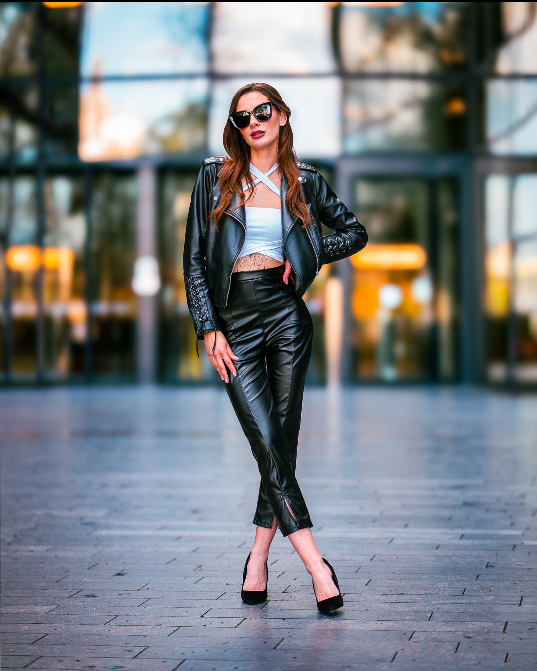 Kobieta w skórzanym stroju i okularach pozuje na tle przeszklonego budynku. Ujęcie pełnej sylwetki, z nogami skrzyżowanymi w kostkach. Lekki uśmiech i pewna postawa.