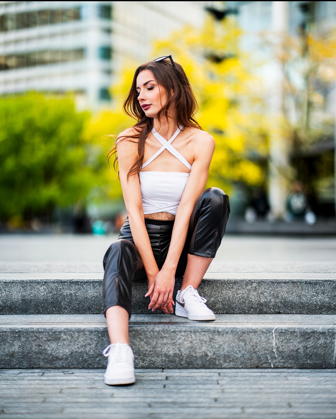 Kobieta siedzi na schodach w białym topie i czarnych spodniach, z budynkiem w tle. Uwagę przyciąga jej poza i miejski krajobraz.