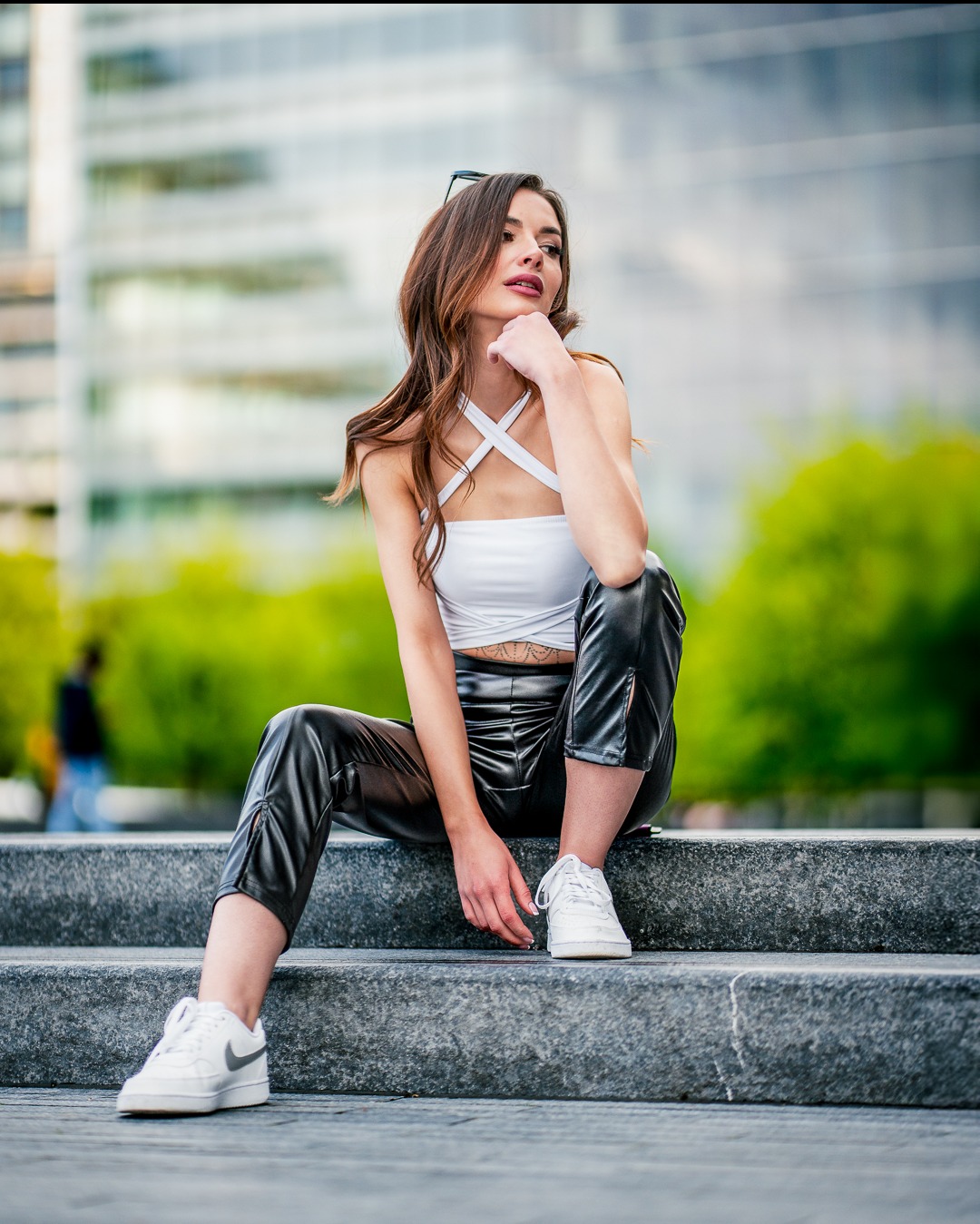 Zamyślona brunetka w białym topie i czarnych spodniach siedzi na schodach, z budynkiem w tle. Sesja w miejskiej scenerii, z delikatnym rozmyciem tła.