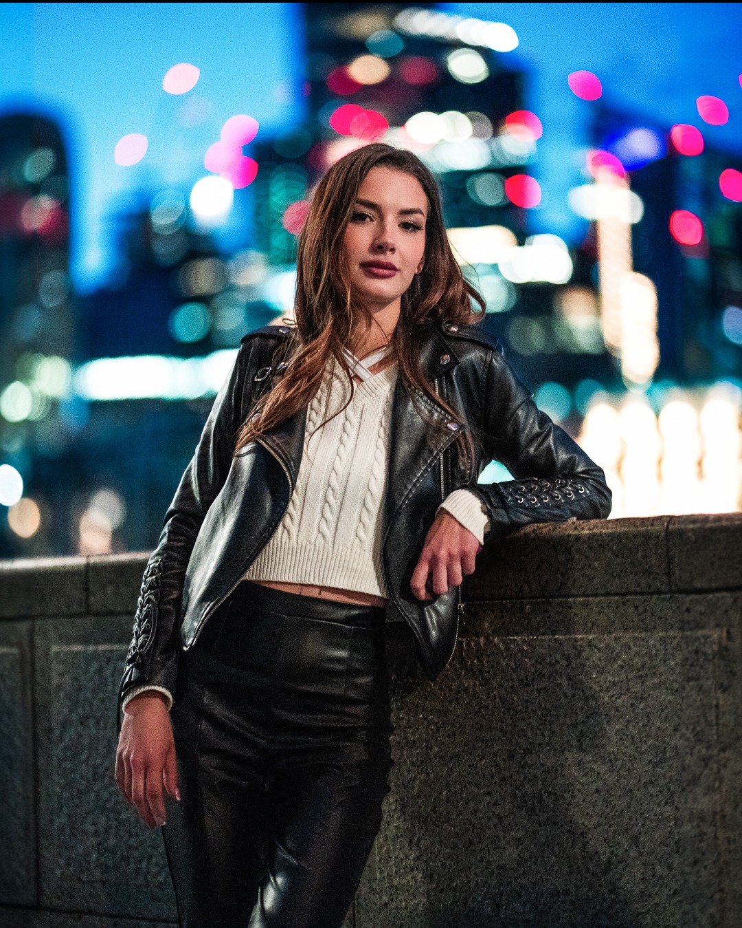 Portret kobiety w skórzanej kurtce na tle nocnego miasta z rozmytymi światłami. Ujęcie z dołu, podkreślające sylwetkę i miejski klimat.