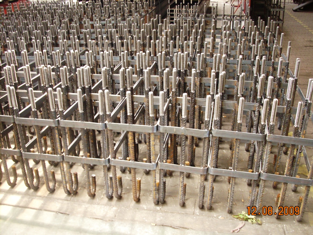 Magazyn wstępnie przygotowanych szkieletów z żebrowanej stali zbrojeniowej, ustawionych pionowo i połączonych płaskownikami, gotowych do zalania betonem.