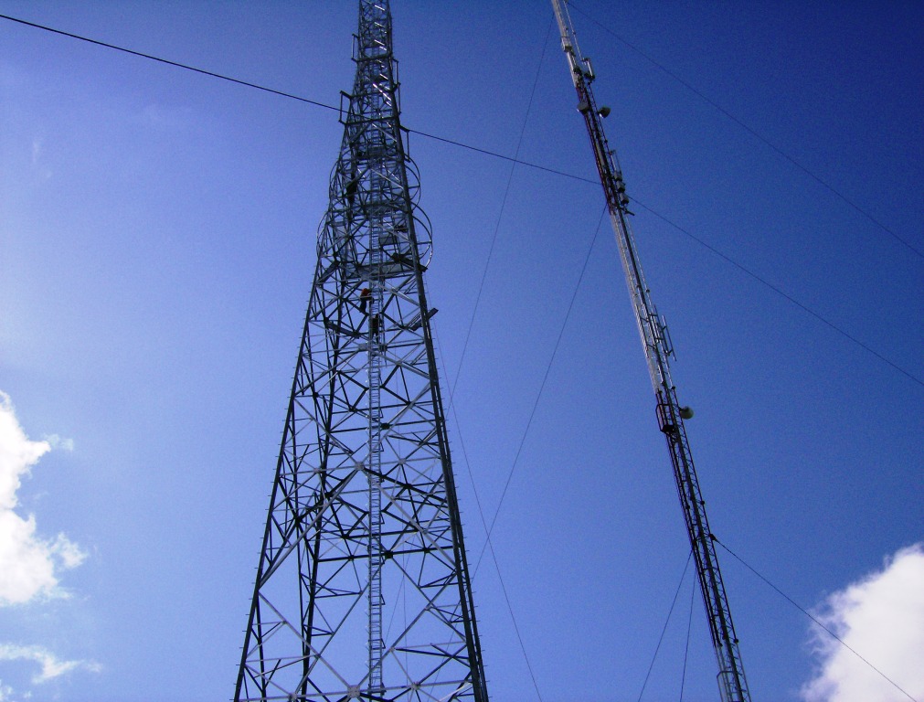 Dwie wieże telekomunikacyjne o stalowej konstrukcji kratownicowej na tle błękitnego nieba z lekkimi obłokami, widok z dołu, jedna z wież z zamontowanymi antenami.