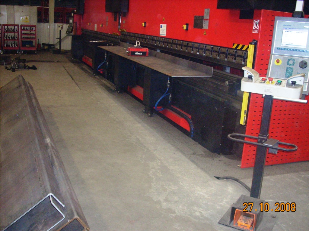 Proces gięcia blachy na dużej, czerwonej prasie krawędziowej w hali produkcyjnej. Widoczny panel sterowania, elementy mocujące i fragment gotowego profilu. Na podłodze widoczna data.