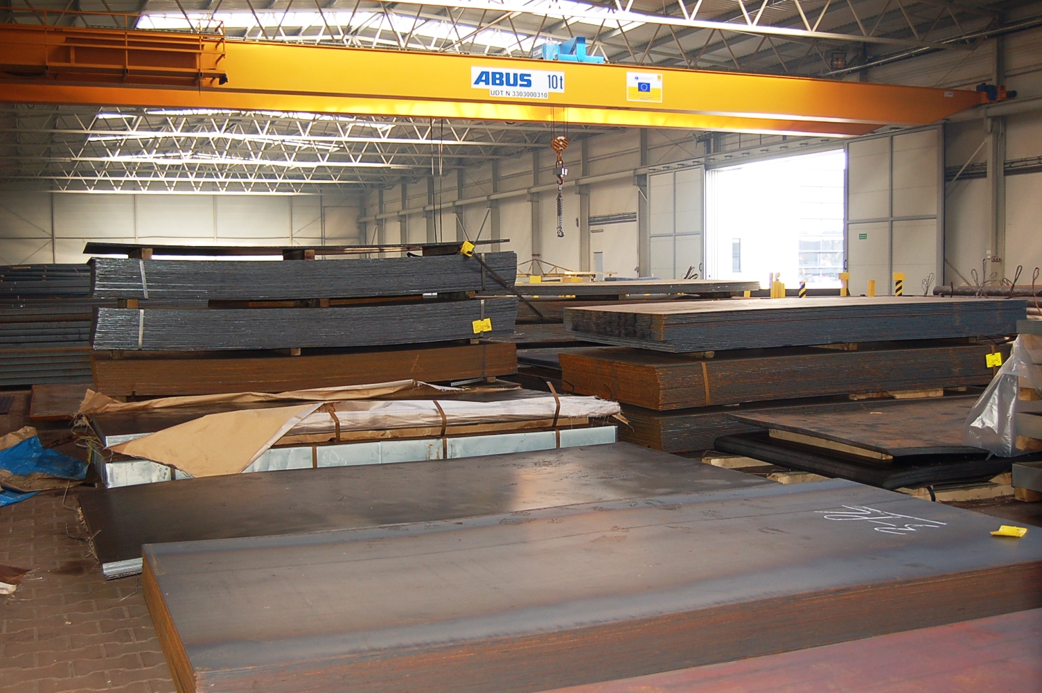 Magazyn wyrobów hutniczych: płaskie i kątowe elementy stalowe ułożone w stosy, widoczny suwnicowy dźwig ABUS 10t pod sufitem hali.