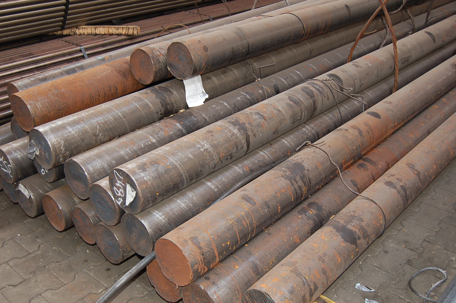 Stalowe pręty o okrągłym przekroju, ułożone warstwami, z widocznymi śladami rdzy i etykietami na niektórych końcach.