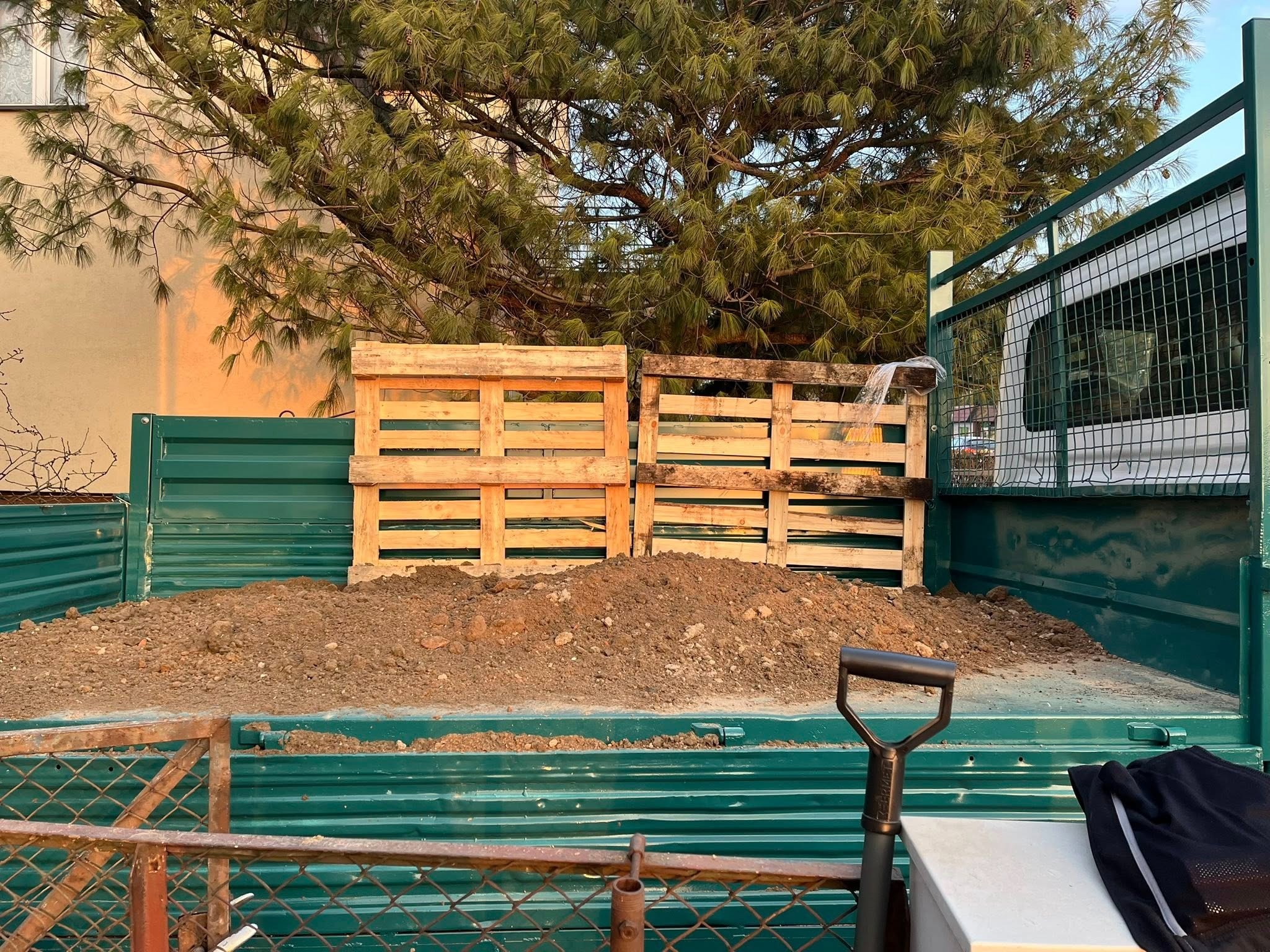 Wnętrze skrzyni ładunkowej zielonego samochodu z ziemią, dwiema paletami i łopatą. W tle widoczne drzewo iglaste i fragment budynku.