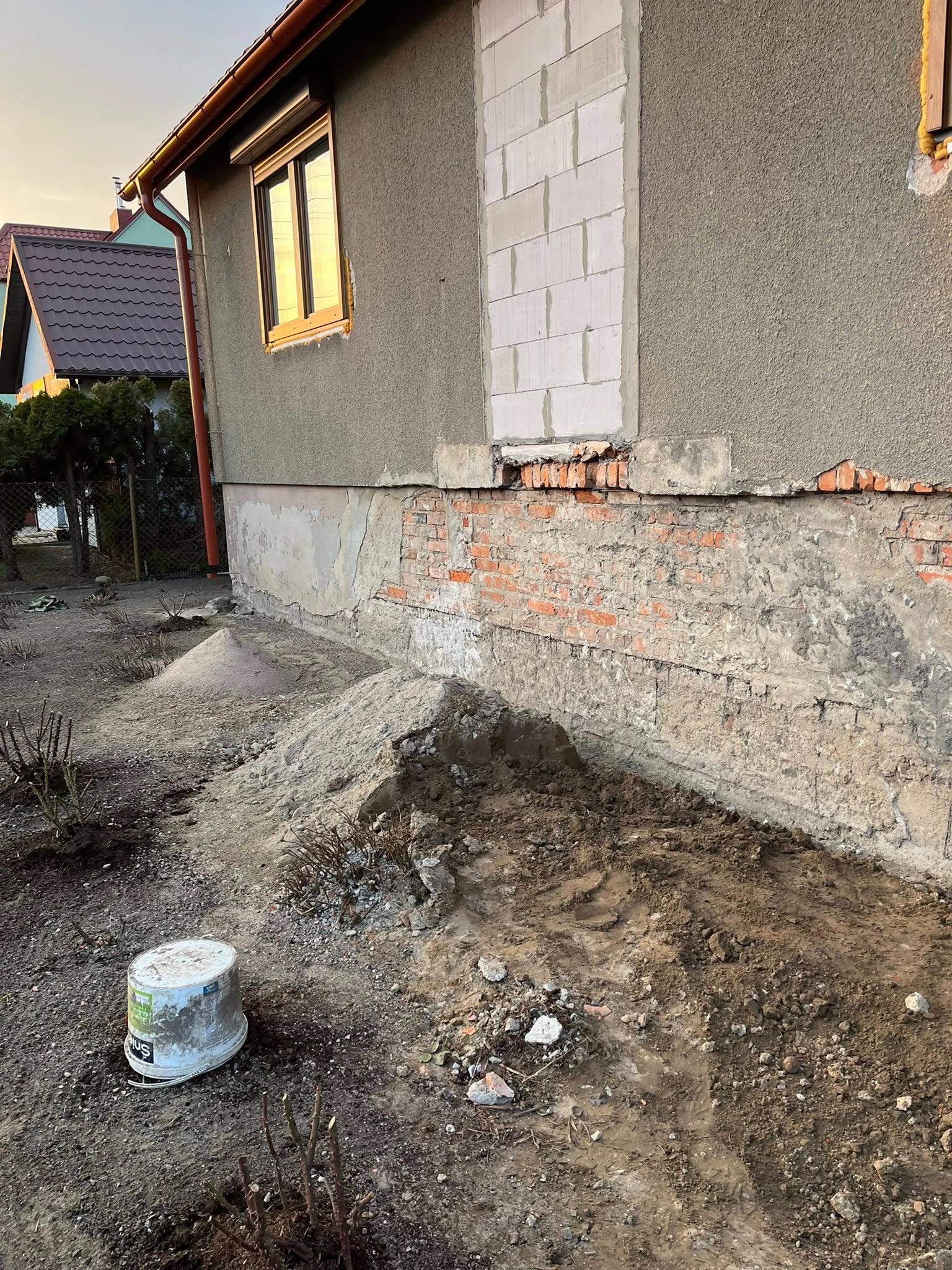 Odkopane fundamenty domu z widoczną cegłą i resztkami tynku, obok pryzma ziemi i wiadro. Fragment elewacji z oknem i pustakami. Prace ziemne przy budynku.