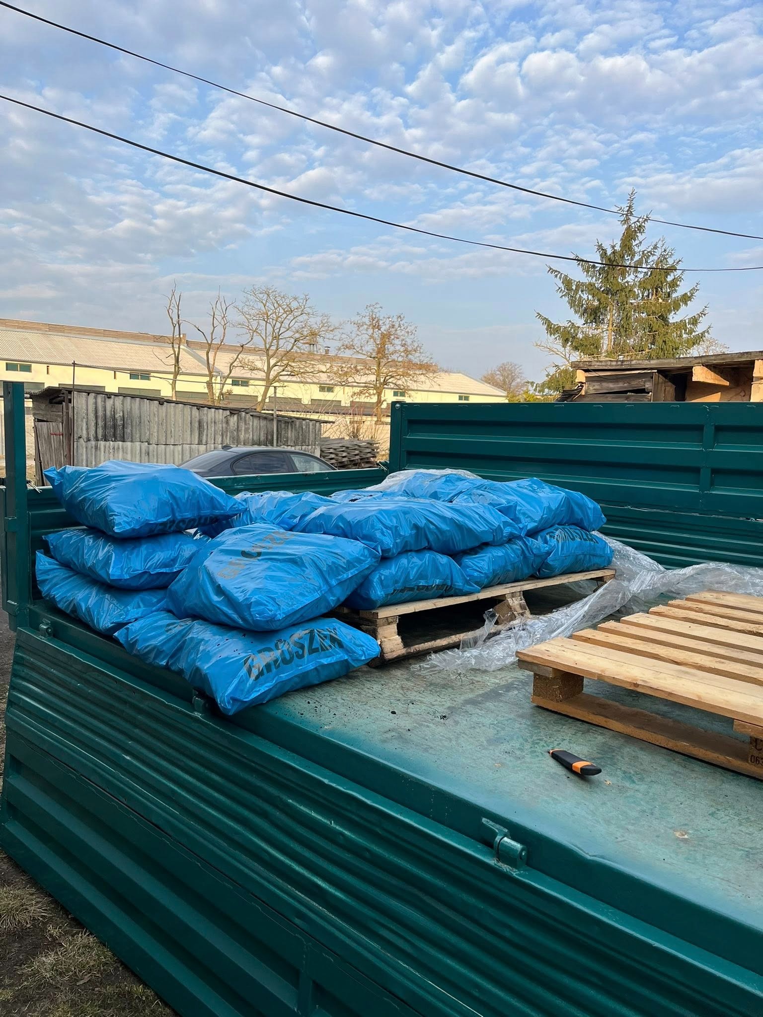 Załadunek worków z gruzem na paletach do zielonej skrzyni samochodu ciężarowego. W tle budynek i drzewo iglaste, błękitne niebo z chmurami.