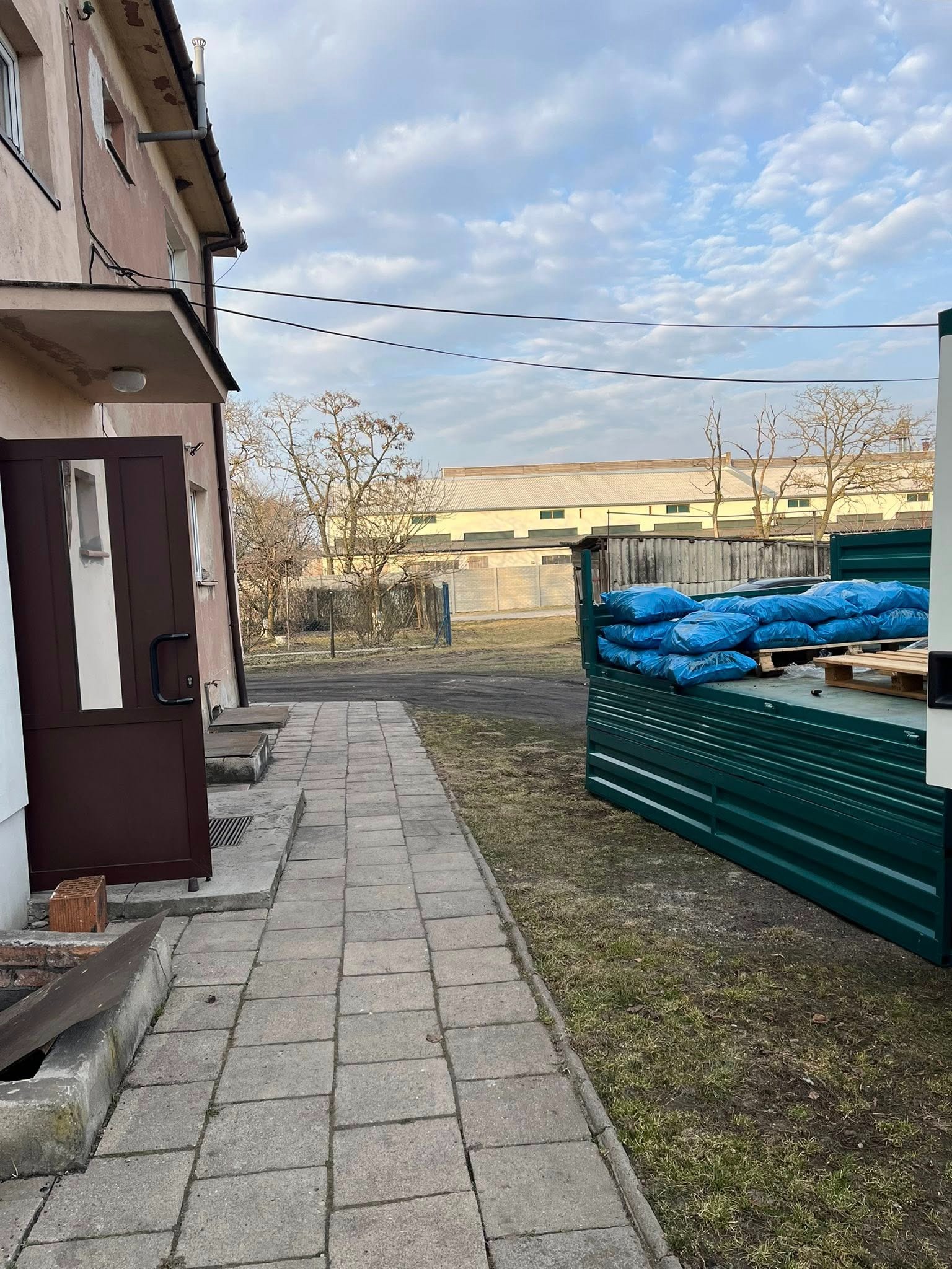 Ciężarówka z niebieskimi workami na pace, zaparkowana obok budynku. Widoczny chodnik z kostki i fragment trawnika. W tle budynek przemysłowy i błękitne niebo.