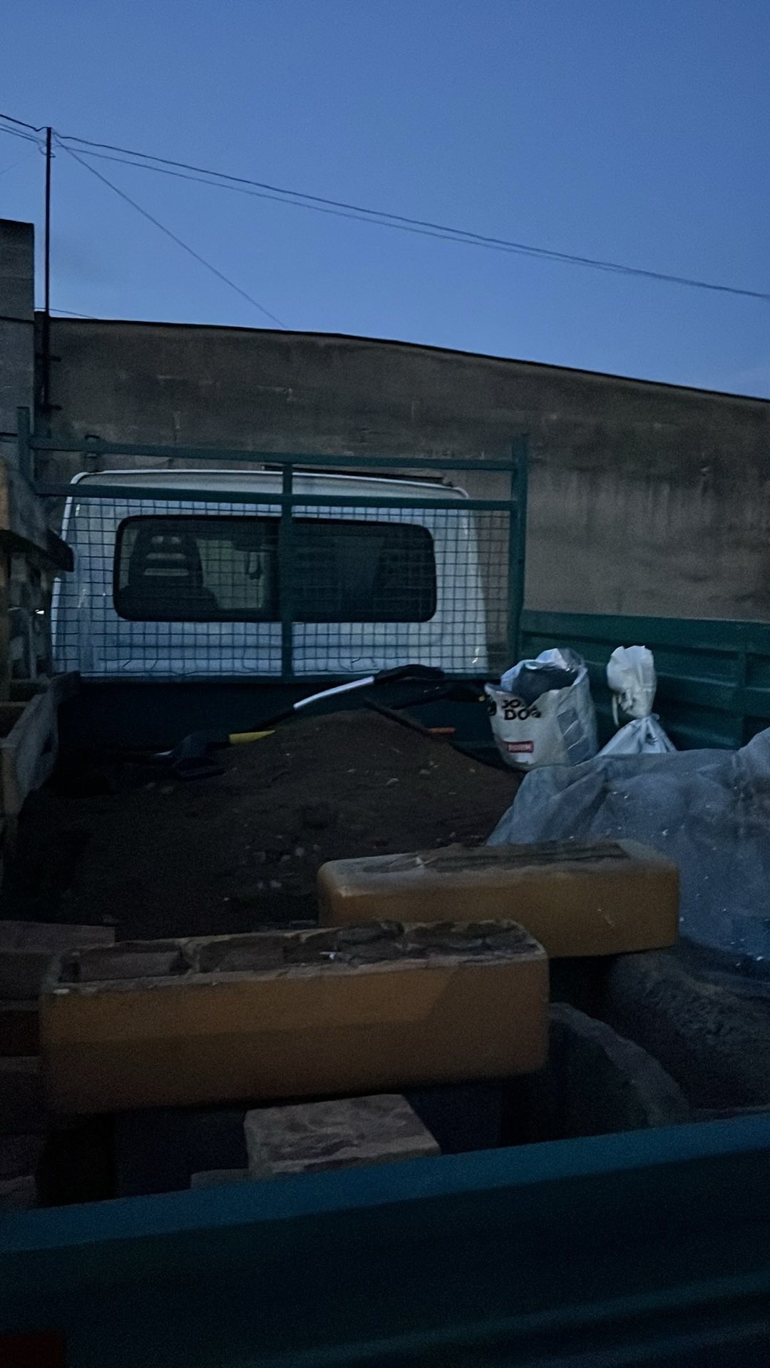 Ciężarówka z otwartą skrzynią ładunkową wypełnioną ziemią, gruzem i workami. Widoczne narzędzia i cegły. Zdjęcie zrobione o zmierzchu.