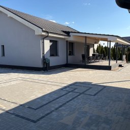 Brukarstwo Adam Drążek - Jasnoszara kostka brukowa ułożona we wzór na podjeździe przed domem z tarasem. Widoczne detale: pergola, meble ogrodowe, schludny ogród. Słoneczny dzień.