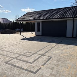 Brukarstwo Adam Drążek - Nowoczesny podjazd z szarej kostki brukowej ułożonej we wzór jodełki, z ciemniejszymi pasami tworzącymi geometryczny deseń. W tle garaż z podcieniem i fragment ogrodu.