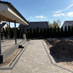 Brukarstwo Adam Drążek - Ułożona kostka brukowa w jodełkę tworzy ścieżkę wzdłuż domu z tarasem. Po prawej stronie widoczna pryzma piasku, a w tle rząd tuj i domy.