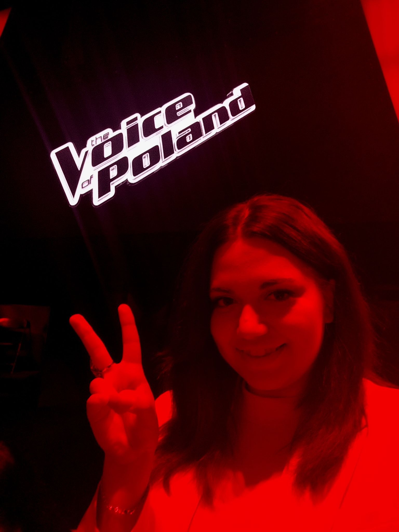 Uśmiechnięta kobieta z symbolem zwycięstwa na tle logo 'The Voice of Poland' w czerwonym oświetleniu. Zdjęcie zza kulis programu talent show.