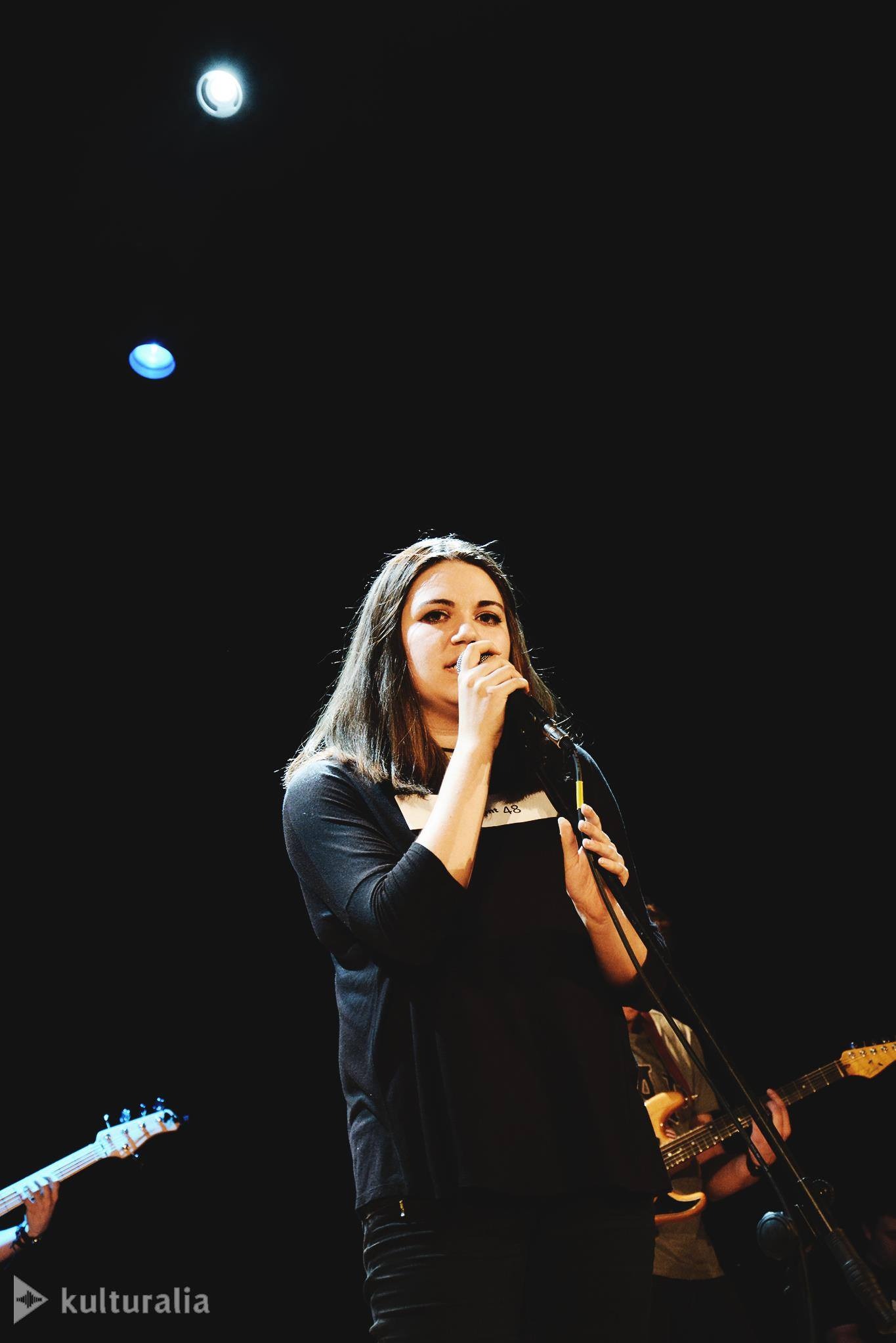 Wokalistka z mikrofonem na scenie, w tle gitary basowa i elektryczna. Scena oświetlona punktowo, ciemne tło. Ujęcie z dołu, widoczny fragment logo 'kulturalia'.