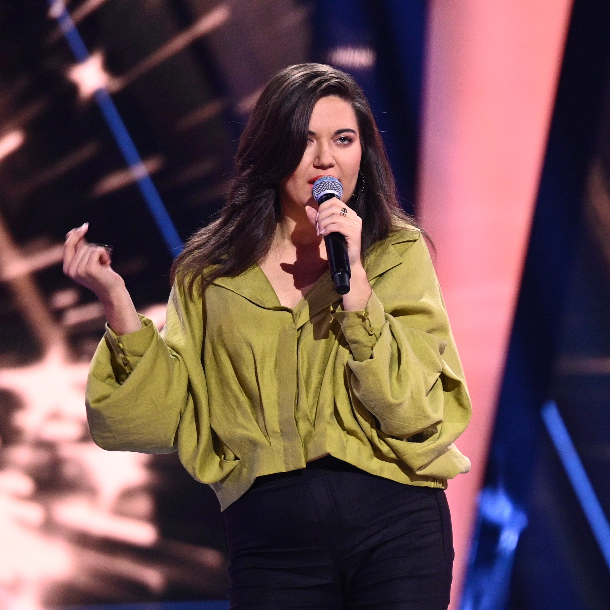 Wokalistka z mikrofonem na scenie, w oliwkowej koszuli i czarnych spodniach, z rozpuszczonymi, ciemnymi włosami, w trakcie występu na tle rozświetlonej sceny.