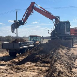 MARK KOP - Pomarańczowa koparka Hitachi załadowuje ziemię do przyczepy ciągniętej przez traktor na tle błękitnego nieba. Widoczne słupy energetyczne i pole.