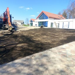 MARK KOP - Niwelacja terenu pod nowoczesny dom z czerwonym dachem. Pomarańczowa koparka na działce, przygotowana pod trawnik. Kostka brukowa w pierwszym planie.