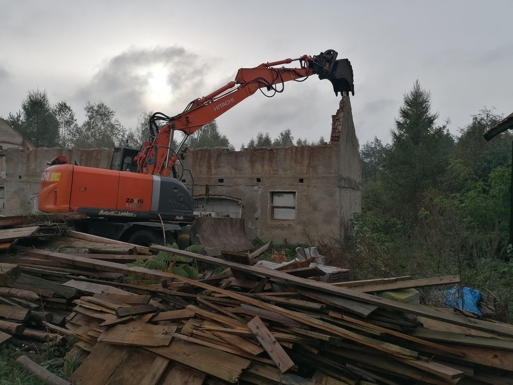 Pomarańczowa koparka Hitachi podczas rozbiórki murowanego budynku w Wydminach. W tle pochmurne niebo i drzewa, w pierwszym planie sterta drewnianych desek.