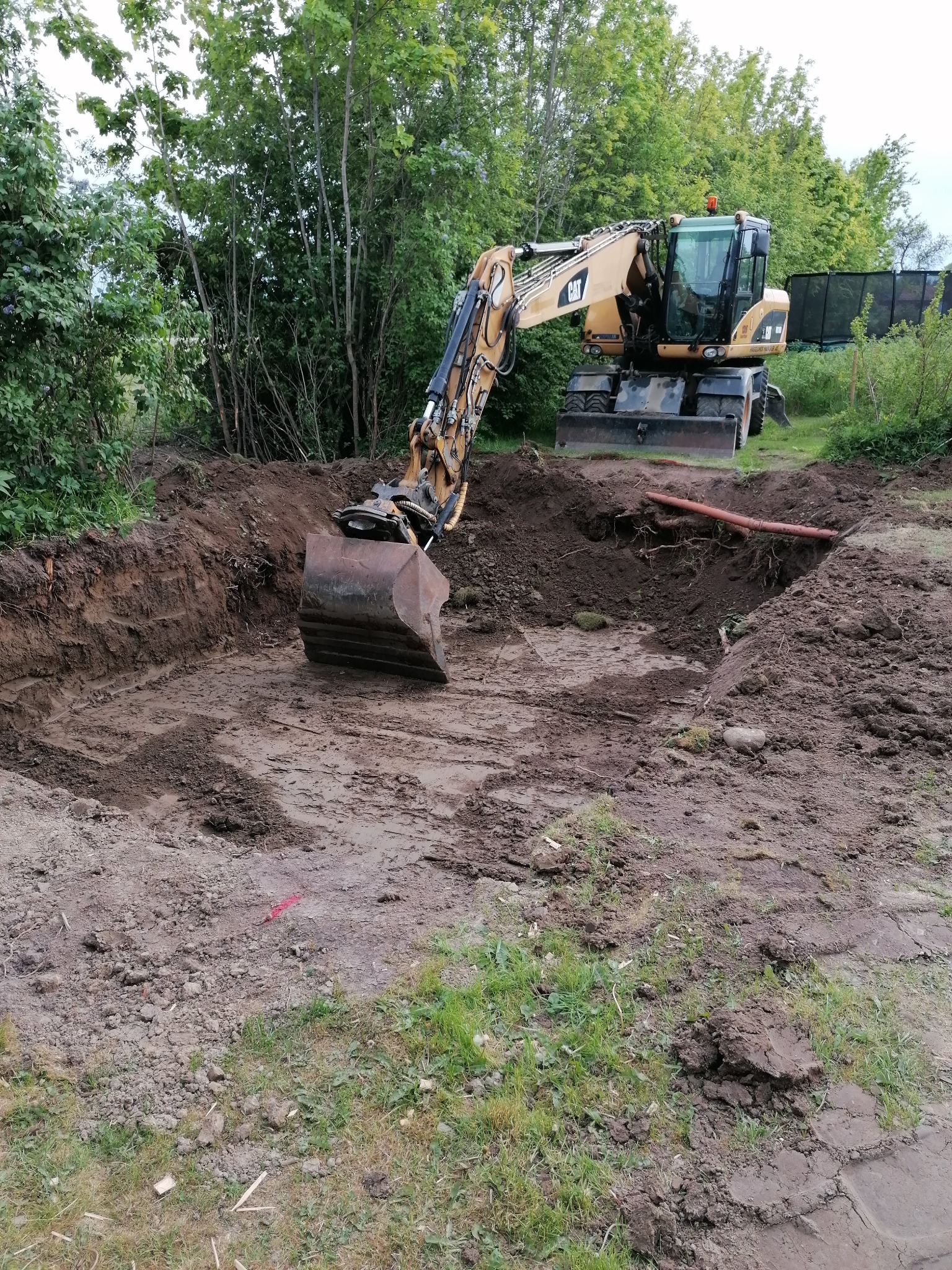 Żółta koparka kołowa CAT w trakcie prac ziemnych, widoczny wykop w gruncie z rurą, otoczony zielenią. Ujęcie z poziomu gruntu.