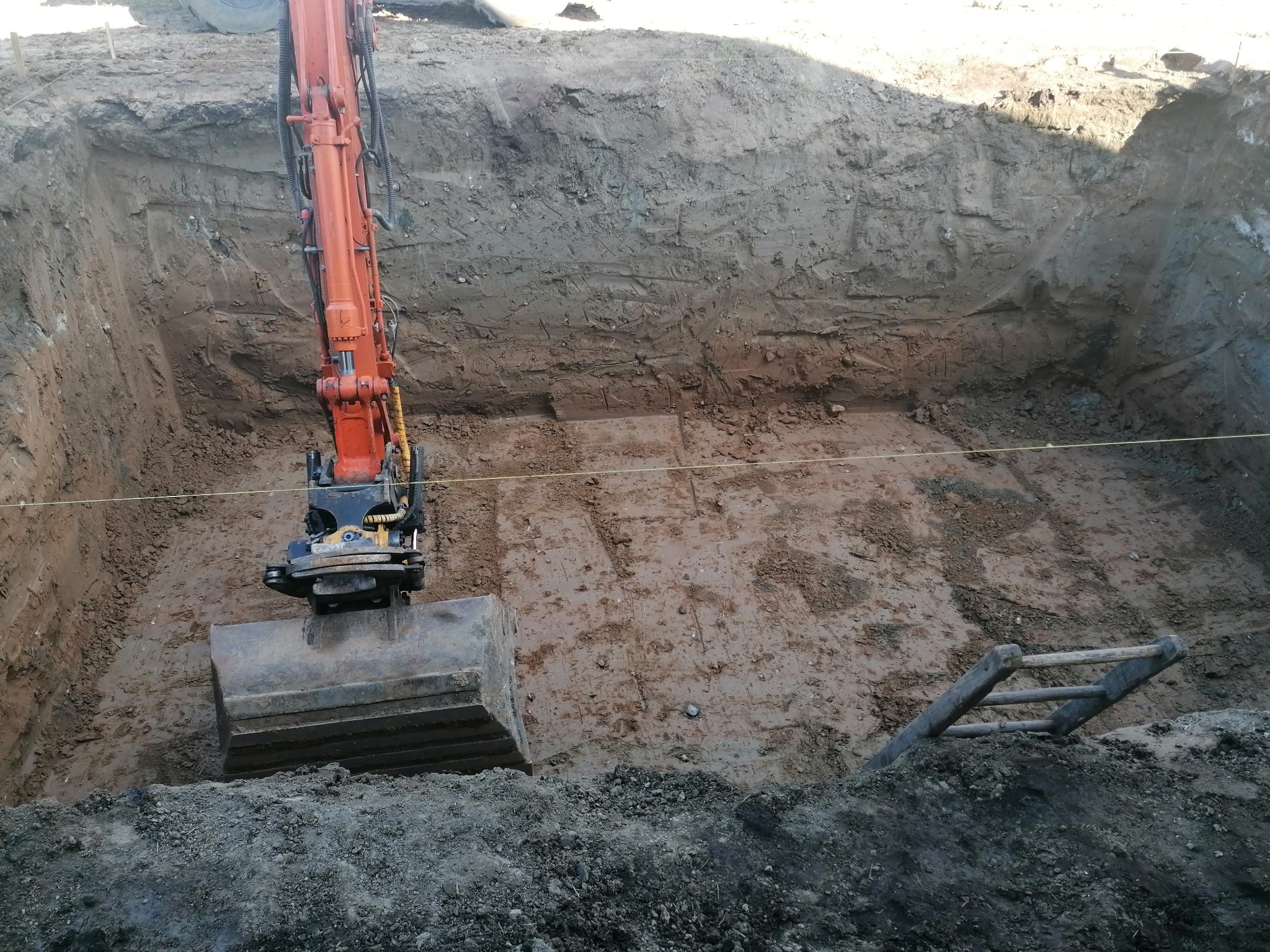Pomarańczowa koparka w głębokim wykopie o nierównych ścianach, z widoczną drabinką i fragmentem opony. W tle ziemia i liny wyznaczające granice.