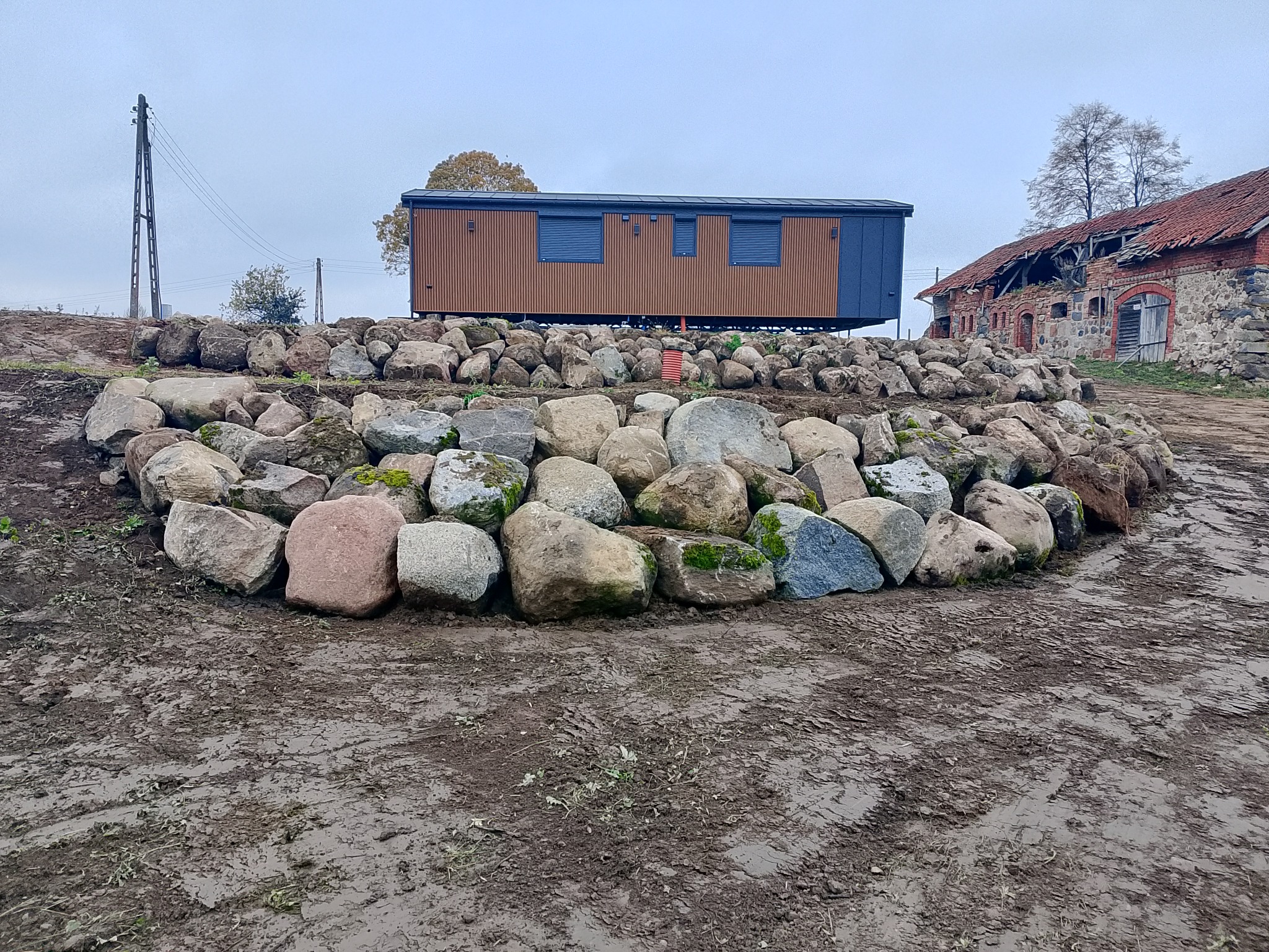 Ustawiony na kamiennym nasypie brązowy kontener biurowy z niebieskimi akcentami, w tle zrujnowany budynek z czerwonej cegły i błotniste podłoże.