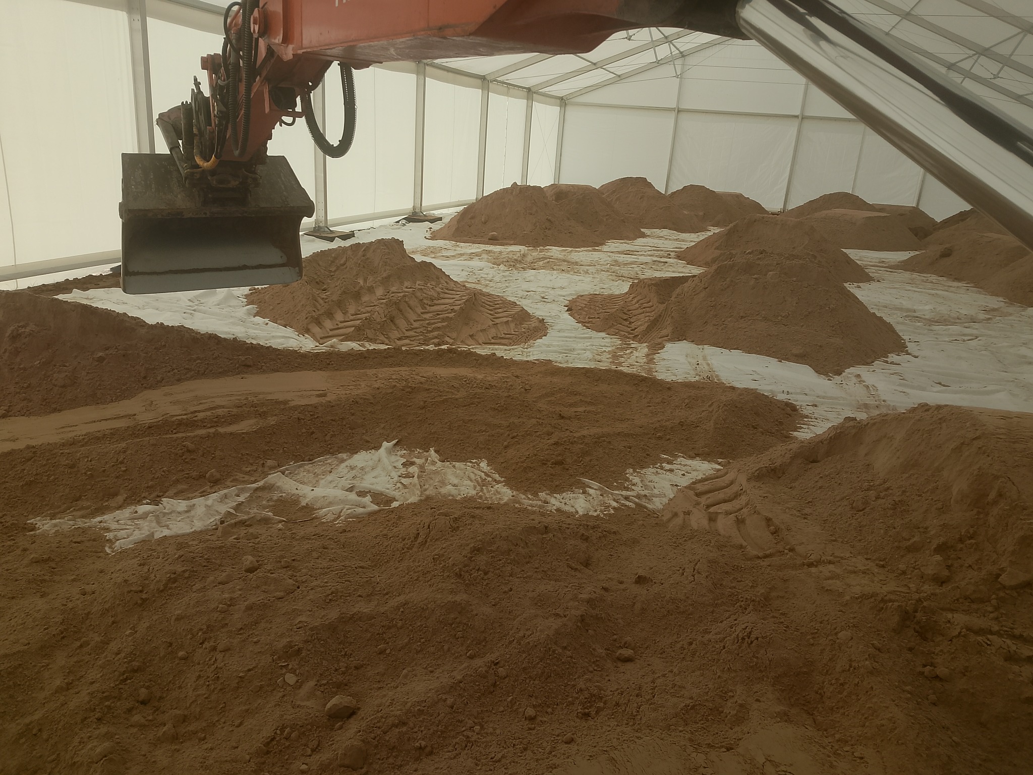 Koparka z łyżką w hali, hałdy ziemi na białej folii. Widoczne ślady opon. Hala z białego materiału, konstrukcja stalowa. Nietypowe miejsce robót ziemnych.
