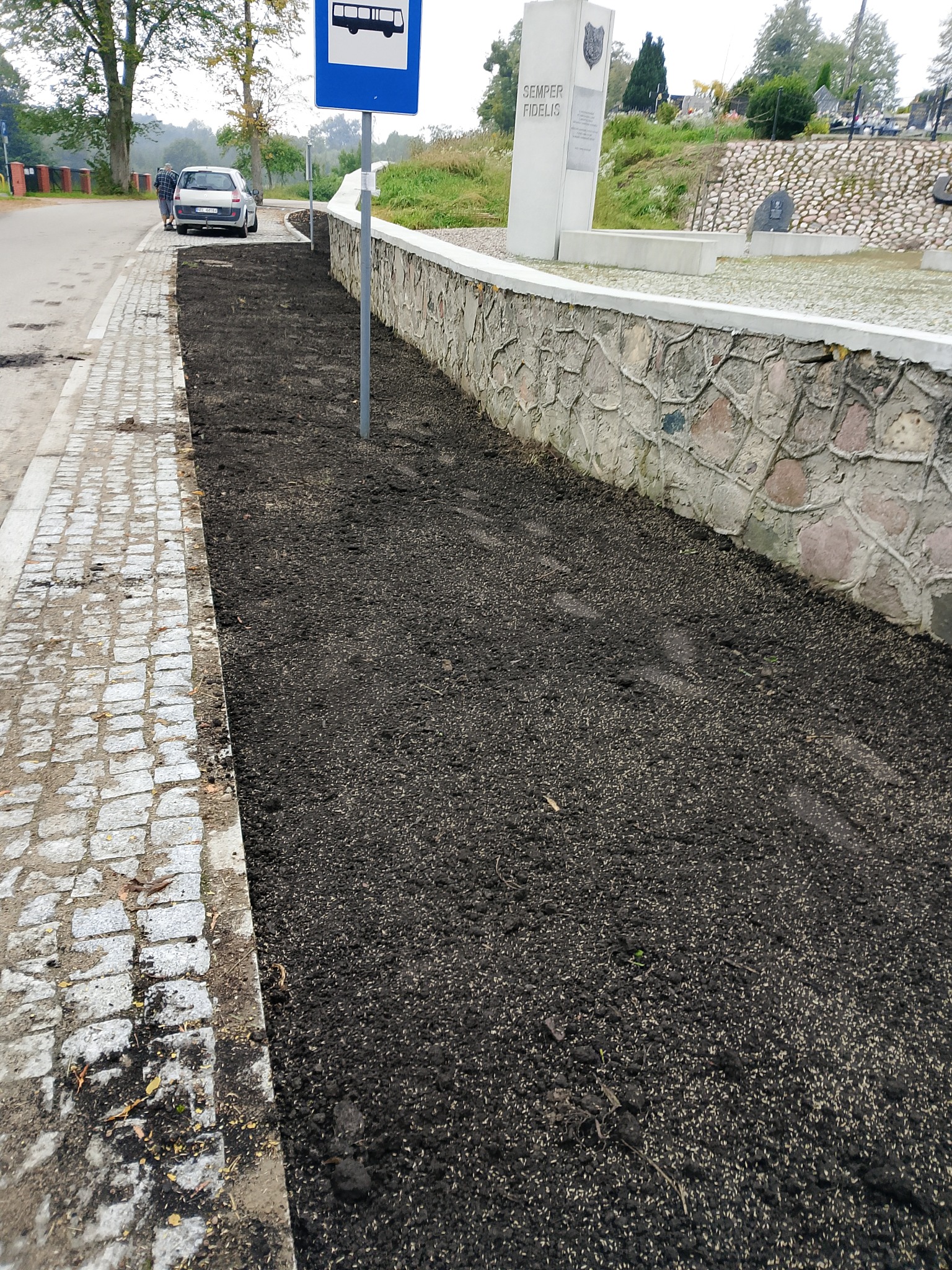 Przygotowany teren pod wysiew trawy wzdłuż kamiennego murku i chodnika z kostki, obok przystanku autobusowego. Widoczne nasiona w ziemi.