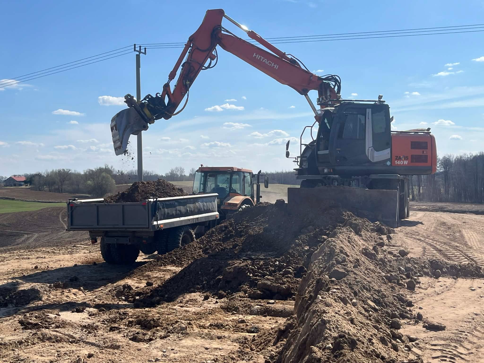 Pomarańczowa koparka Hitachi załadowuje ziemię do przyczepy ciągniętej przez traktor na tle błękitnego nieba. Widoczne słupy energetyczne i pole.