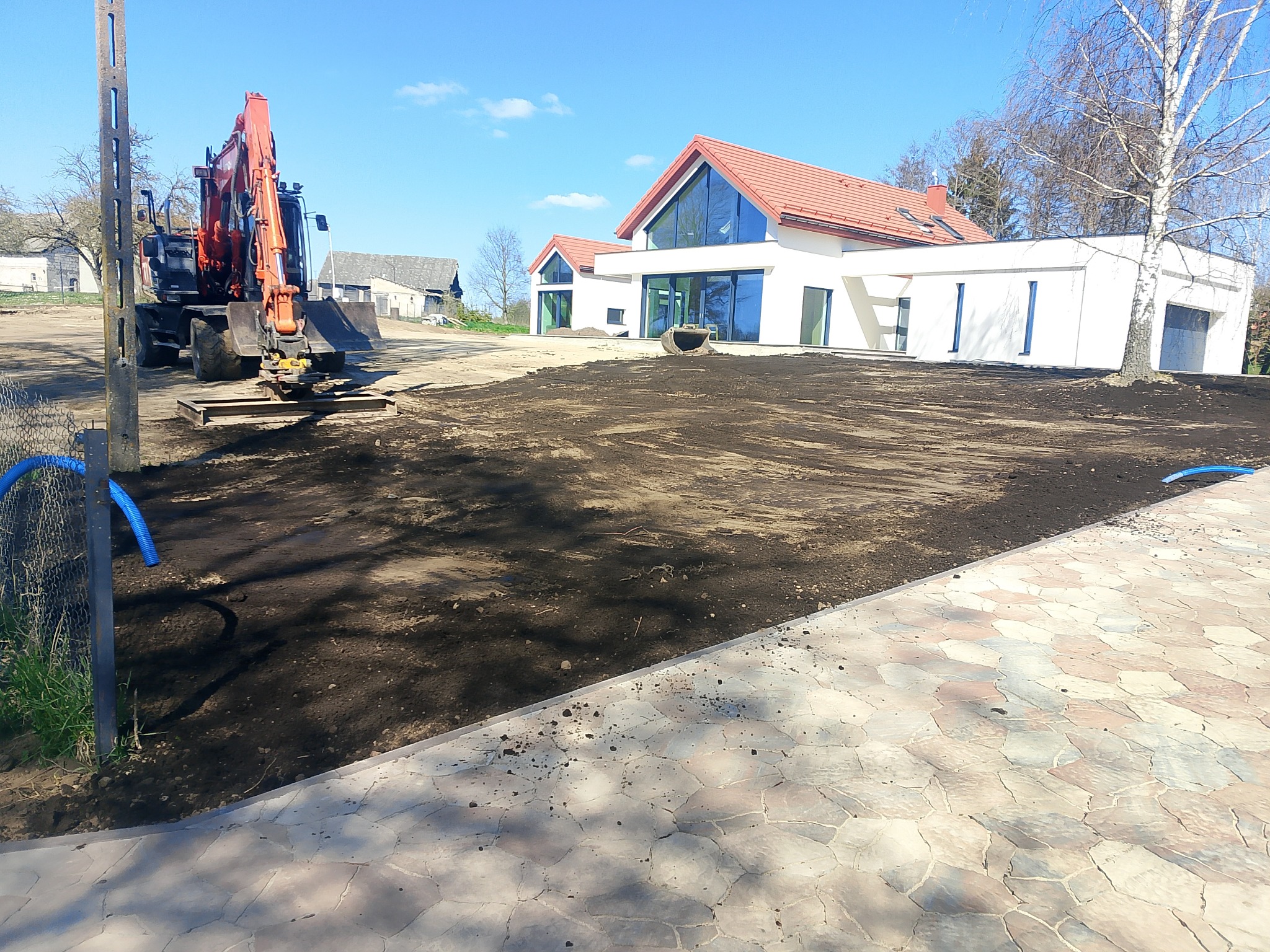 Niwelacja terenu pod nowoczesny dom z czerwonym dachem. Pomarańczowa koparka na działce, przygotowana pod trawnik. Kostka brukowa w pierwszym planie.
