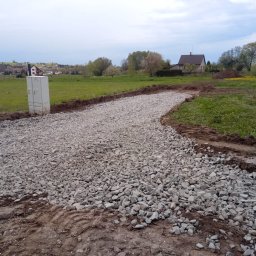 KDK Budownictwo Grzegorz Żydek - Utwardzona żwirowa droga gruntowa na tle zielonego krajobrazu z domami w oddali. Widoczna skrzynka elektryczna i ślady opon. Pochmurne niebo.