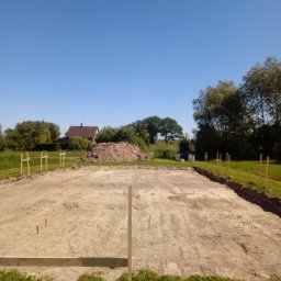 KDK Budownictwo Grzegorz Żydek - Wytyczony teren pod budowę domu z drewnianymi słupkami i palikami. W tle dom z brązowym dachem i sterta ziemi. Błękitne niebo.