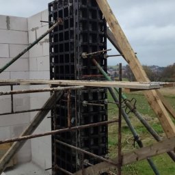 KDK Budownictwo Grzegorz Żydek - Szczegółowy widok szalunku kolumny żelbetowej zbrojonej stalą, wspartego drewnianymi belkami i metalowymi stemplami, na tle muru z bloczków i zieleni.