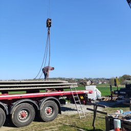 KDK Budownictwo Grzegorz Żydek - Pracownik zabezpiecza płyty żelbetowe na naczepie ciężarówki, używając łańcucha z hakiem dźwigu. W tle widoczny krajobraz wiejski i błękitne niebo.