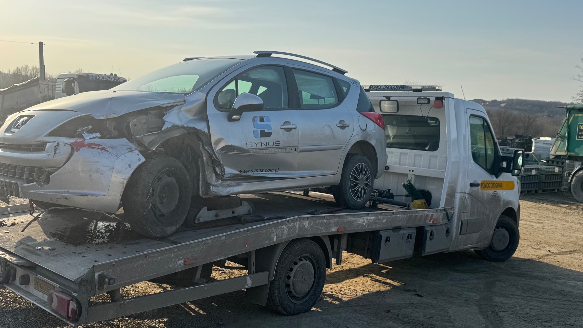 Uszkodzony srebrny Peugeot na platformie białej ciężarówki pomocy drogowej z napisem 'Pomoc Drogowa' i logo firmy Synos, w tle krajobraz.