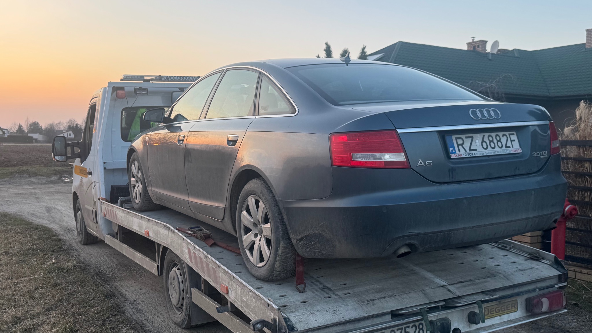 Szary samochód osobowy marki Audi A6 na platformie pomocy drogowej, widok z tyłu, tablica rejestracyjna RZ 6882F, w tle dom i zachodzące słońce.