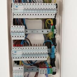 Rozdzielnica elektryczna z widocznymi bezpiecznikami i kolorowymi przewodami, zamontowana w białej obudowie. Ujęcie z bliska, ukazujące szczegóły instalacji.