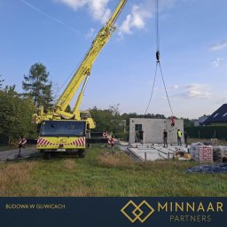 MINNAAR PARTNERS SPÓŁKA Z OGRANICZONĄ ODPOWIEDZIALNOŚCIĄ - Montaż prefabrykowanej ściany domu z pomocą dźwigu. Widoczni pracownicy w kaskach i kamizelkach odblaskowych. Na pierwszym planie fundamenty i materiały budowlane.