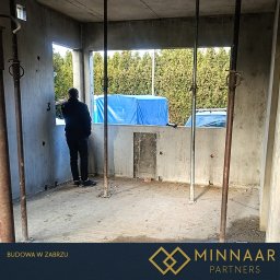 MINNAAR PARTNERS SPÓŁKA Z OGRANICZONĄ ODPOWIEDZIALNOŚCIĄ - Surowy, betonowy szkielet budynku w trakcie budowy. Widoczne podpory, otwory okienne i fragment sylwetki osoby obserwującej otoczenie. W tle zieleń i niebieska plandeka.