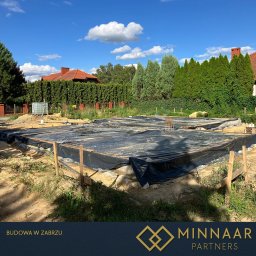 MINNAAR PARTNERS SPÓŁKA Z OGRANICZONĄ ODPOWIEDZIALNOŚCIĄ - Przygotowany szalunek pod fundamenty domu jednorodzinnego, zabezpieczony czarną folią. Widoczne zbrojenia. W tle zieleń drzew i błękitne niebo.